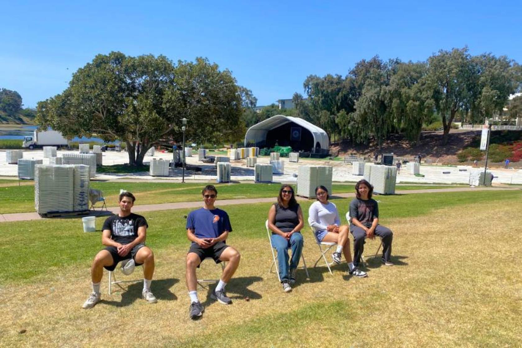4 student workers sitting in chairs on a grass lawn with commencement set-up in the background